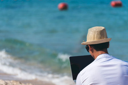 Man traveler use laptop on summer beach blue sky. Asian man wear casual short pants tropical shirt style relax on the beach in summertime. Man typing laptop computer  holiday business tripの写真素材