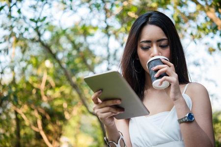 Asian Woman use smartphone drinking coffee hand holding hot disposable cup in green park. Banner...