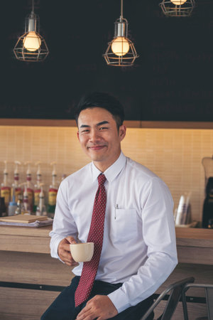 Asian Businessman drinking coffee cup relax working at office desk. CEO Manager men holding coffee cup work in cozy office. Close up man hand holding black Tea cup smile happiness face at coffee shopの写真素材
