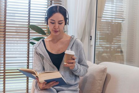 Happy asian woman use headphones listen to music reading book drink coffee cup sitting on cozy sofa...