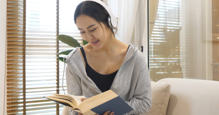 Happy asian woman use headphones listen to music reading book sitting on cozy sofa in living room at...