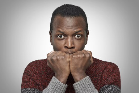 Portrait of scared young African American man dressed in sweater keeping hands in fists, holding them in front of his face, looking at camera with shocked and frightened expression. Fear concept.の写真素材