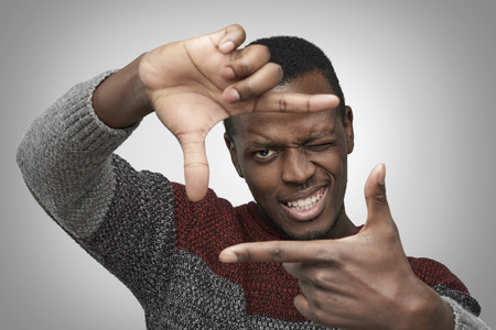 Close up portrait of young African American male in warm casual sweater making frame with his hands, looking at camera and smiling, standing against isolated grey background. Dark-skinned photographerの写真素材