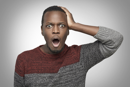 Portrain of terrified young African American man in casual sweater. holding hand on his head and screaming with horrified look, opening mouth widely. Black male feeling scared or stressed. OMG conceptの写真素材