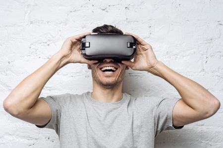 Astonished handsome guy in gray t-shirt experiencing virtual reality while using oculus rift headset for entertaining, standing against white brick wall. People, technology and innovation concept. の写真素材