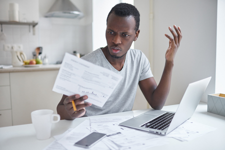 Indignant angry irritated african american male sitting at kitchen table, looking at papers in shock, astonished with amount of unpaid bills. Financial stress, feeling depressed and frustratedの写真素材