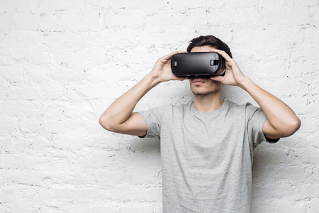 Calm and confident handsome european male wearing grey t-shirt and virtual reality headset or 3d glasses, playing video game or watching movie against white brick wall. Technology, innovation conceptの写真素材