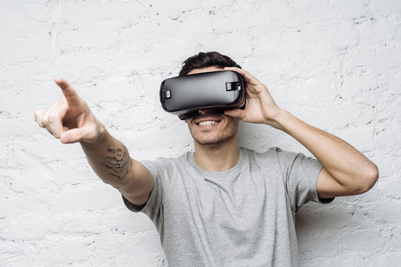 Smiling man using oculus rift headset, experiencing virtual reality while playing video game. Young tattooed european male wearing 3d glasses, pointing his finger as if interacting with something の写真素材