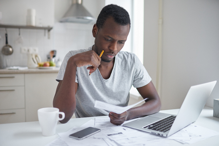 Stressed african american man working through paperwork, calculating expenses, trying to save some money, managing finances, sitting at kitchen table with laptop, trying to make finance planの写真素材