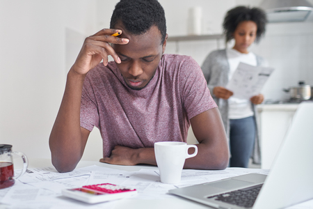 Depressed bankrupt young african man feeling financial stress while reading notification, informing about eviction from their house because of non-payment for rent. Wife reading bills on background の写真素材