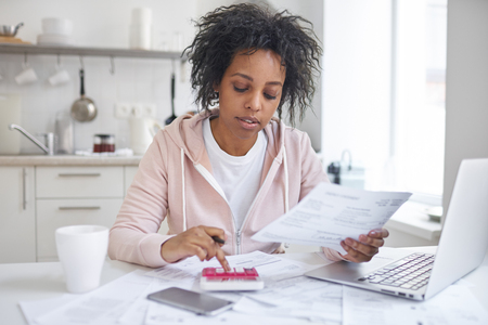 Financial literacy concept. Indoor shot of young single african american female student sitting at kitchen table, calculating expences, trying to analyze domestic dudget, thinking how to pay debtの写真素材