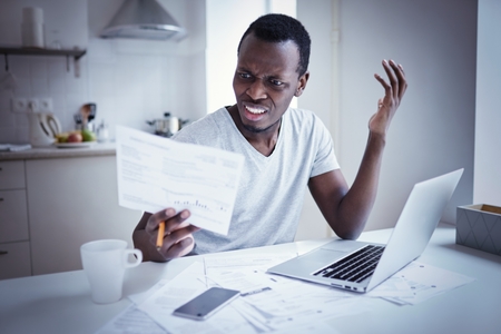 Indignant angry irritated young african american man looking at utility bill, frustrated about high taxes, worried about troubles with mortgage payment to bank, having serious problems with debtの写真素材