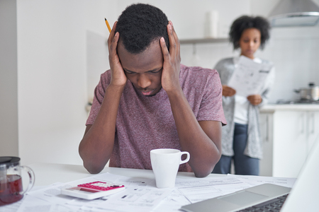African male sitting at kitchen table, feeling sad and depressed about debts, can't pay off his utility bills. Wife standing in background with papers from bank. People and finances conceptの写真素材