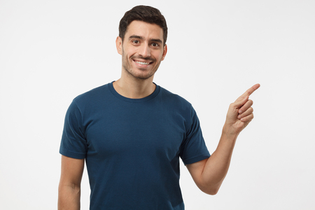 Casual dressed young man in blue t-shirt pointing right with his finger isolated on gray backgroundの写真素材