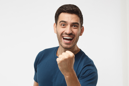 Indoor photo of good-looking male isolated on gray background celebrating victory and acting as if he is winner, squeezing fists in deep emotional expression of happiness and luckの写真素材