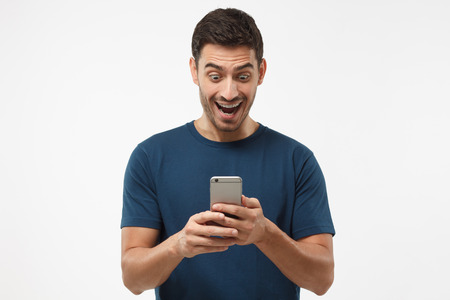Close up shot portrait of young man isolated on gray background, looking agitated at display of her smartphone smiling and laughing happily, impressed by media content from webの写真素材