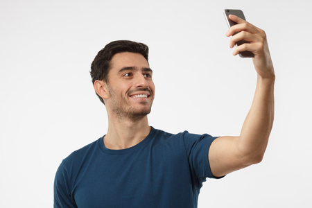 Portrait of young handsome caucasian man standing against gray background using smartphone to take selfie pictures and smilingの写真素材
