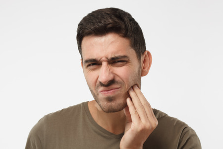 Indoor headshot of young male feeling pain, holding his cheek with hand, suffering from bad toothache, looking at camera with painful expression. Tooth ache concept.の写真素材