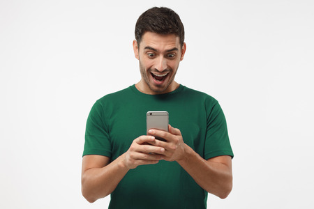 Close up portrait of student isolated on gray background, looking agitated at display of his smartphone smiling and laughing happily, impressed by media content from webの写真素材