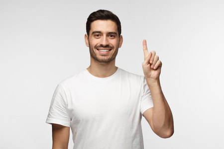 Attractive young man in white t-shirt pointing up with his finger isolated on gray backgroundの写真素材