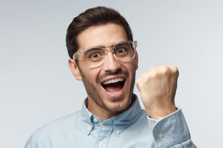 Studio portrait of young handsome European man pictured isolated on gray background with transparent safety glasses showing fist with positive face expression as if having achieved his goalの写真素材