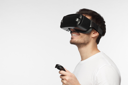 Closeup headshot of young handsome European man pictured isolated on gray background wearing virtual reality headset and smiling while pressing button on remote control he is holding in handの写真素材