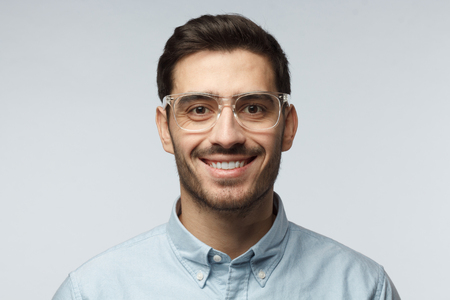 Close up shot of smiling attractive business man in blue shirt and trendy trasparent eyeglasses isolated on gray backgroundの写真素材