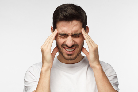 Portrait of young man isolated on grey background suffering from severe headache, pressing fingers to temples, closing eyes to relieve pain with helpless face expressionの写真素材