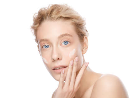Young beautiful blonde woman with clear skin applying face cream with fingers, isolated on whiteの写真素材