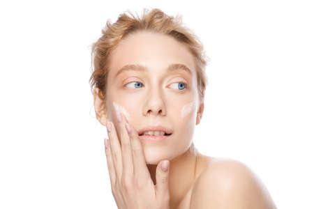 Beautiful blonde woman with glowing skin applying face cream with fingers, isolated on whiteの写真素材