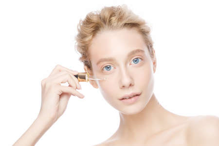 Young woman with blue eyes and blond hair applying serum to her face, isolated on whiteの写真素材