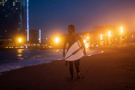 Night shot of young surfer holding surf board and walking along the coast, ready for surfingの写真素材