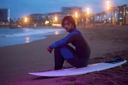 Night portrait of surfer in neoprene suit sitting on surf board and looking at camera after surfingの写真素材