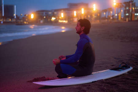 Night portrait of surfer sitting on his surf board and meditating in lotus pose after surfingの写真素材