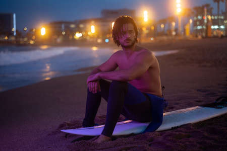 Night portrait of young surfer sitting on his surf board and looking away after surfingの写真素材