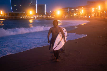 Night shot of fit surfer holding surf board and walking along the coast, ready for surfingの写真素材