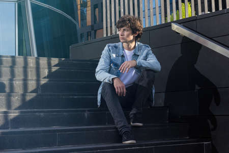 Attractive young guy in jeans t-shirt sits on stairs against business centerの写真素材