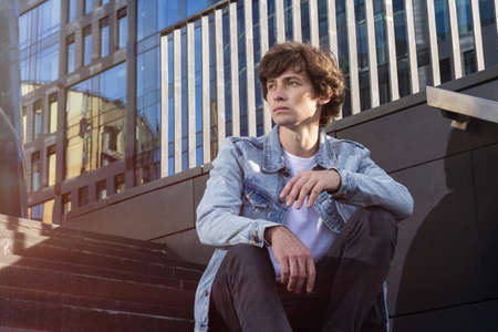 Active casual man in blue jeans jacket sits on stairs against city backgroundの写真素材