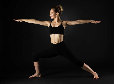Slim well fit caucasian girl doing yoga Warrior pose on dark black backgroundの写真素材
