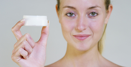 Portrait of a beautiful young woman smiling, looking at camera, without makeup, holding cream, on a white background. Concept: natural beauty, youth, skin care, always young, love yourself.の写真素材