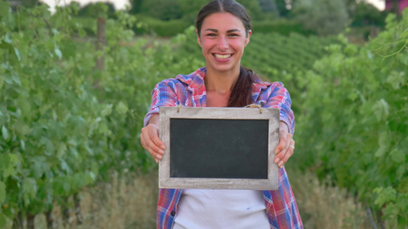 Beautiful girl (woman) farmer smiling watching the grape fields, holding a black board, in a shirt, greens background Concept ecology wine bio product, inspection, water, natural products, agricultureの写真素材