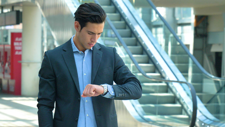 Portrait of a handsome young man in suit, at the station, looking at the clock, smiling. Concept: new business, business relationships, career growth, success.の写真素材
