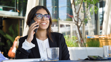 Portrait of a beautiful young business woman (student) in a suit, glasses, sitting at a table, talking on the phone, drinking coffee. Concept: new business, communication, Arab, banker, manager, call.の写真素材