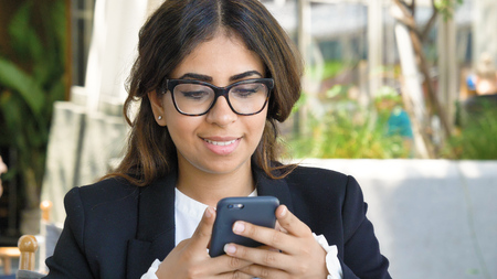 Portrait of a beautiful young business woman (student) in a suit, glasses, sitting at a table, using the phone, drinking coffee. Concept: new business, communication, Arab, banker, manager, call.の写真素材