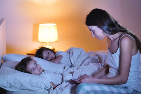 Mom reads a fairy tale of fantasy to her daughters in bed to make her fall asleep and let him have quiet dreams. Concept of: fantasy, relaxation, education, books.の写真素材