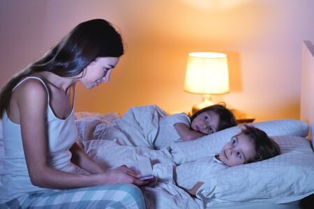 Mom reads a fairy tale of fantasy to her daughters in bed to make her fall asleep and let him have quiet dreams. Concept of: fantasy, relaxation, education, books.の写真素材