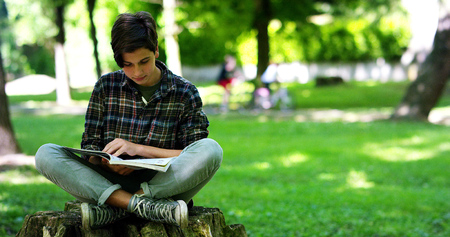Happy young student boy, on a sunny day at the park, uses a tablet to studyの写真素材
