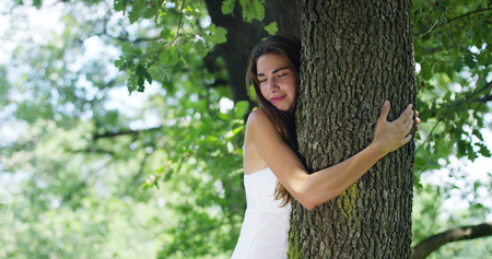 A young woman in a white dress embraces a tree and smiles in a sunny day in a green woods. Concept of freedom, liberty, lifestyle, equilibrium, connection to a nature and a green world, peaceの写真素材