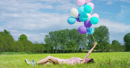 The young woman in pink shovels dress happy and carefree lying on a meadow with colorful balloons in her hands. Concept of happiness and freedomの写真素材