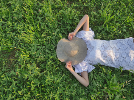 On a sunny day, a woman in a white dress lies on the green grass and hides her face with a straw hat Concept life style ecology colorful happy people freedom wonderfully beautiful, greens, environmentの写真素材